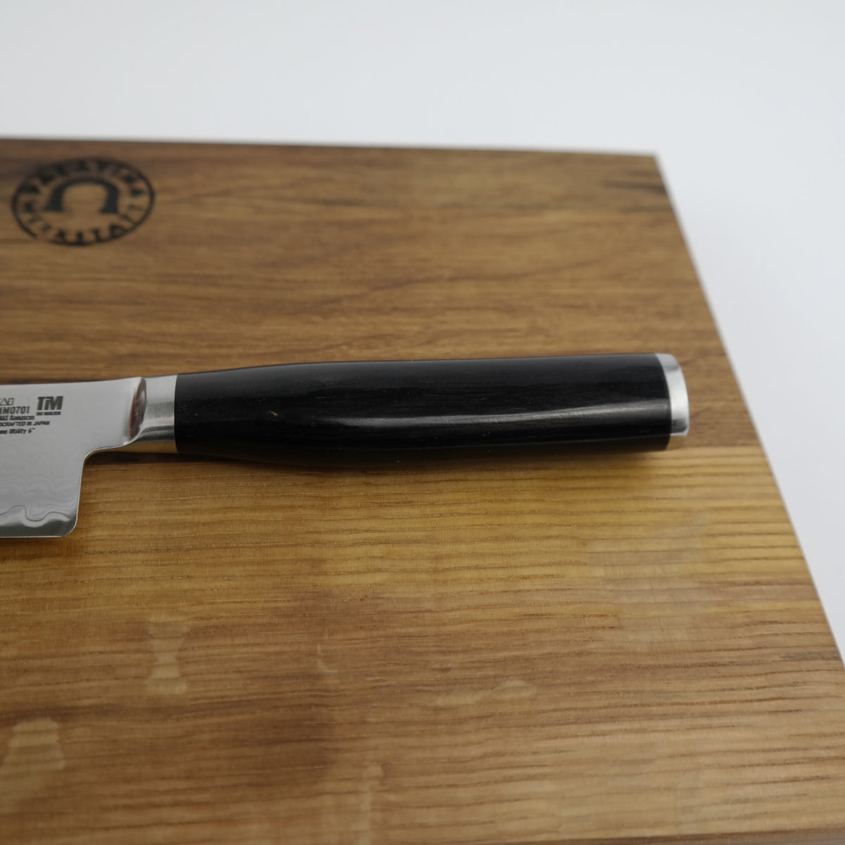 Ein Kai Shun Premier Tim Mälzer Minamo TMM-0701 ultrascharfes Universalmesser mit schwarzem Griff und 15 cm langer Edelstahlklinge liegt auf einem Holzschneidebrett aus alter Fasseiche (25x15 cm). Das Schneidebrett hat in der oberen linken Ecke ein eingestanztes Logo. Die Klinge ist teilweise sichtbar, so dass nur der obere Teil des Messers sichtbar ist.