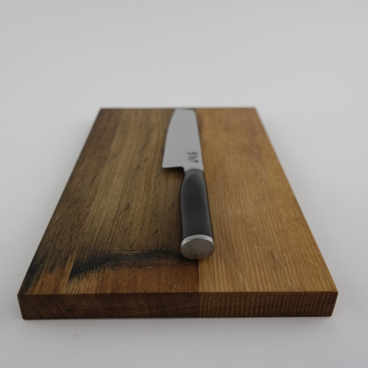 Ein Kai Shun Premier Tim Mälzer Minamo TMM-0701, ein ultrascharfes Allzweckmesser mit einer 15 cm langen Klinge, ruht diagonal auf einem Schneidebrett aus gealtertem Eichenfassholz mit unterschiedlichen Holzmaserungen auf einer weißen Oberfläche.