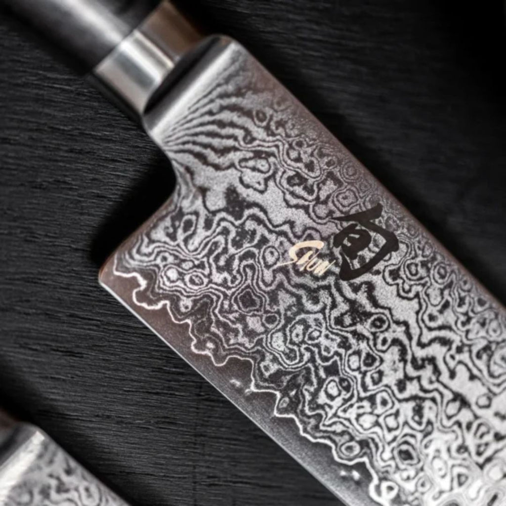 Nahaufnahme des KAI Shun Tim Mälzer Minamo TMM-0702 Santoku, 20cm "THE MAGIC KAI WISHLIST", mit einer Klinge aus Damaszenerstahl mit wirbelnden Mustern und japanischem Kanji-Logo, die auf einer dunklen, strukturierten Oberfläche liegt.