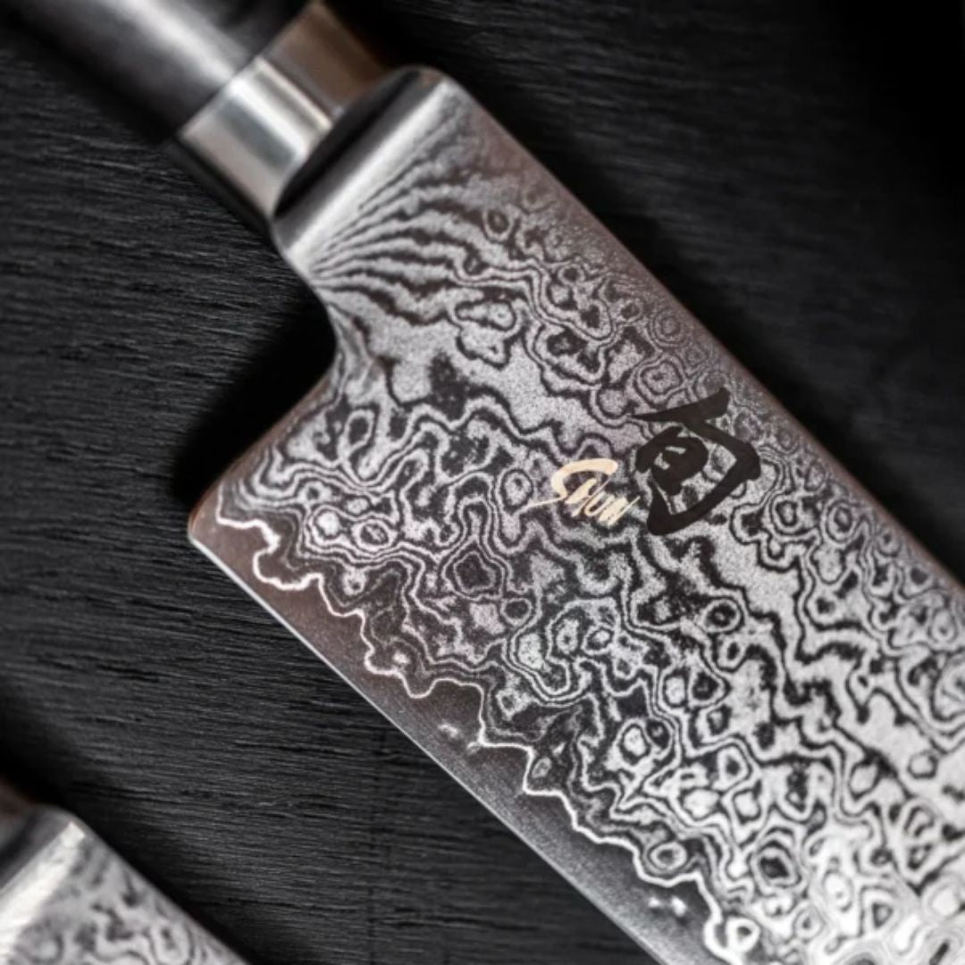 Nahaufnahme des KAI Shun Tim Mälzer Minamo TMM-0702 Santoku, 20cm "THE MAGIC KAI WISHLIST", mit einer Klinge aus Damaszenerstahl mit wirbelnden Mustern und japanischem Kanji-Logo, die auf einer dunklen, strukturierten Oberfläche liegt.