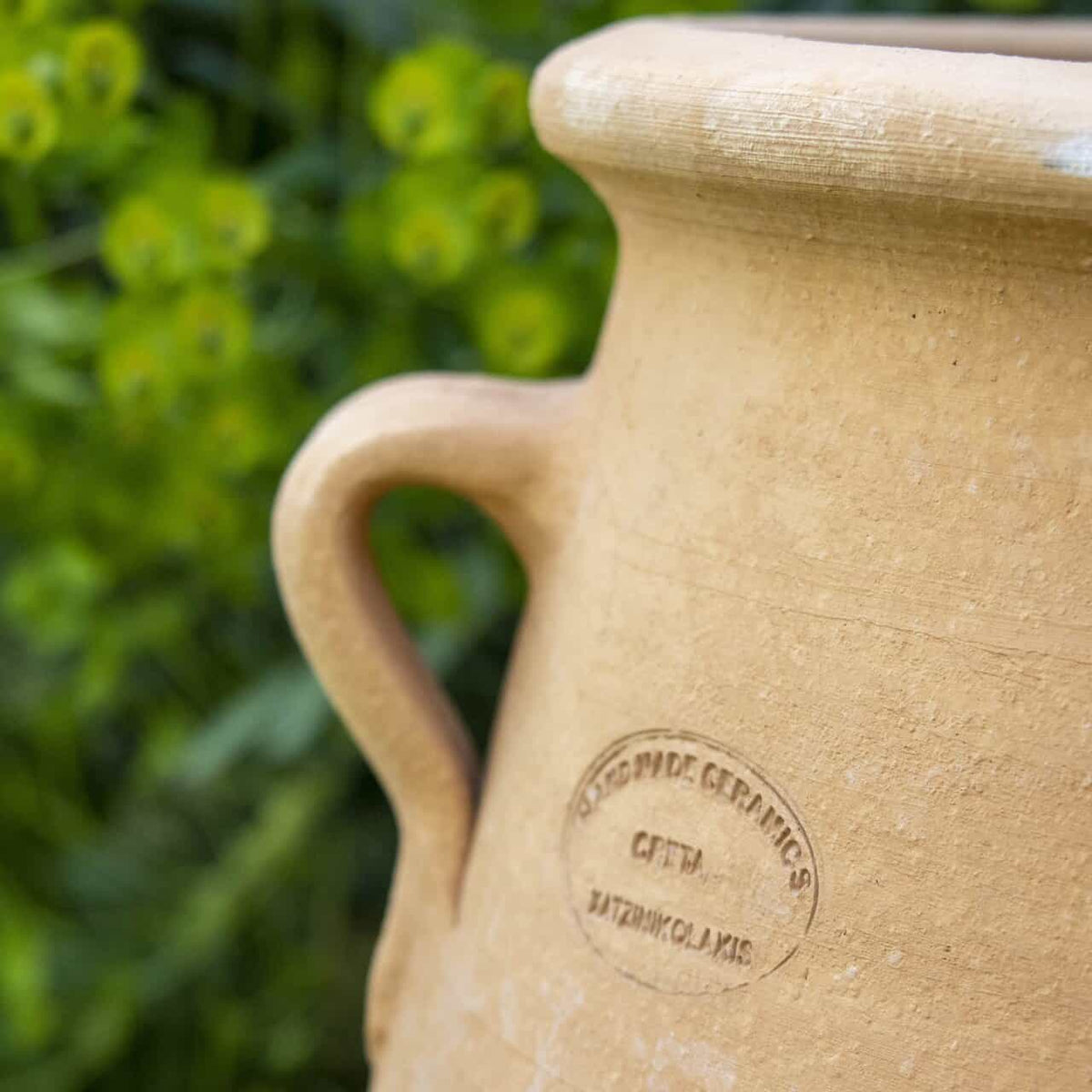 Eine Nahaufnahme der Amphore zum Bepflanzen "Thymus" (H=40cm), ein hellbrauner Keramiktopf mit Henkel und den Prägungen "WORLDWIDE CERAMICS" und "GFITA BATZINIKOALS", inmitten von verschwommenem grünem Laub.