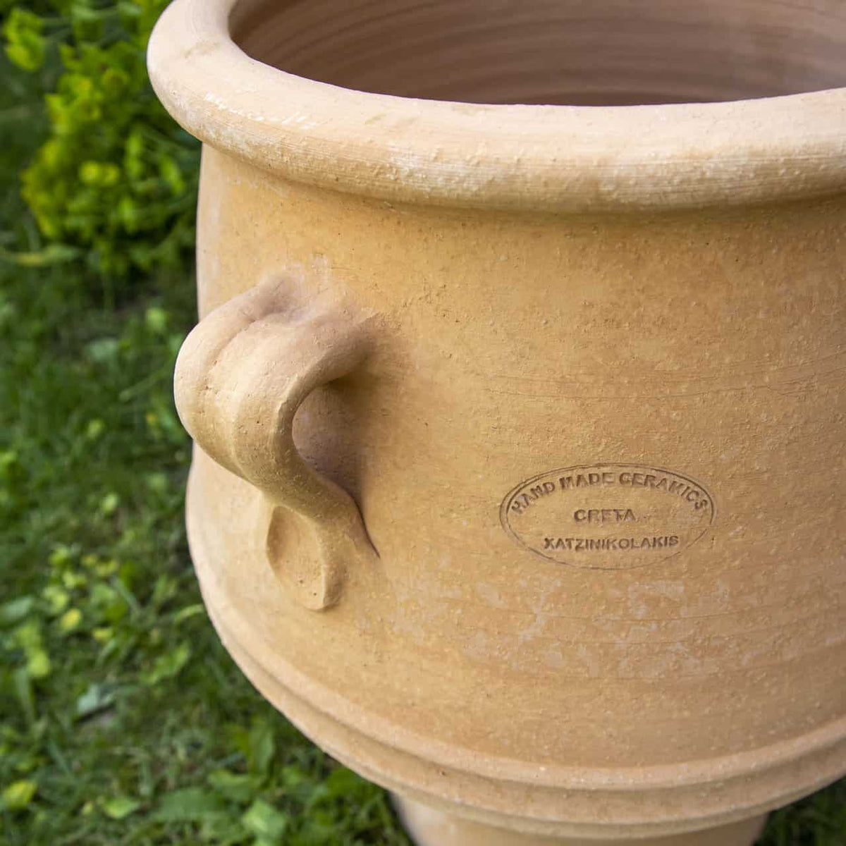 Nahaufnahme der handgefertigten beigen Keramik "Amphore zum Bepflanzen 'Thymus'" (H=40cm) mit geschwungenem Griff und eingraviertem Etikett, platziert auf grünem Gras zwischen Blattpflanzen und Thymus im Hintergrund.