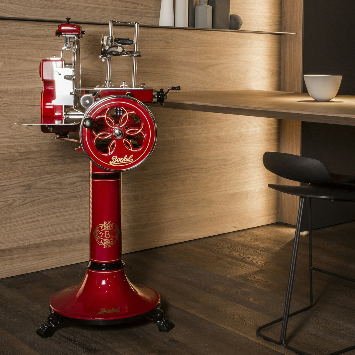 Eine Vintage-Aufschnittmaschine Berkel Volano Tribute Rot mit Blumenschwungrad steht neben einer modernen Holztheke mit schlichtem, elegantem Design. Die Aufschnittmaschine mit ihren filigranen Griffen und der glänzenden Oberfläche bildet einen Kontrast zum minimalistischen Dekor, zu dem ein schwarzer Stuhl und eine weiße Schüssel auf der Theke gehören. Abgerundet wird das Ganze durch ein Aufschnittbrett aus Fassholz, das neben der Aufschnittmaschine steht.
