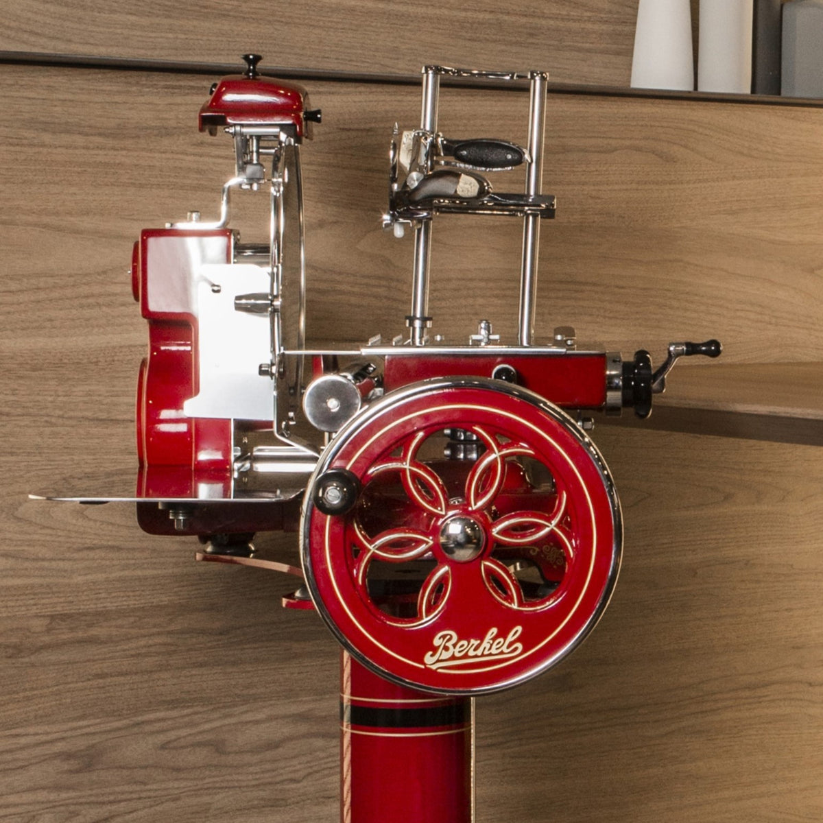 Ein roter, klassischer Berkel Volano Tribute Rot-Fleischschneider mit polierten Metallkomponenten und eleganten Designdetails. Der Schneider verfügt über ein großes Blumenschwungrad-Handkurbelrad, das mit einem komplizierten Muster verziert ist, und den Markennamen „Berkel“ in Kursivschrift in der Nähe seiner Basis. Der Hintergrund ist eine Holzoberfläche, die aus Weinfassdauben gefertigt wurde.