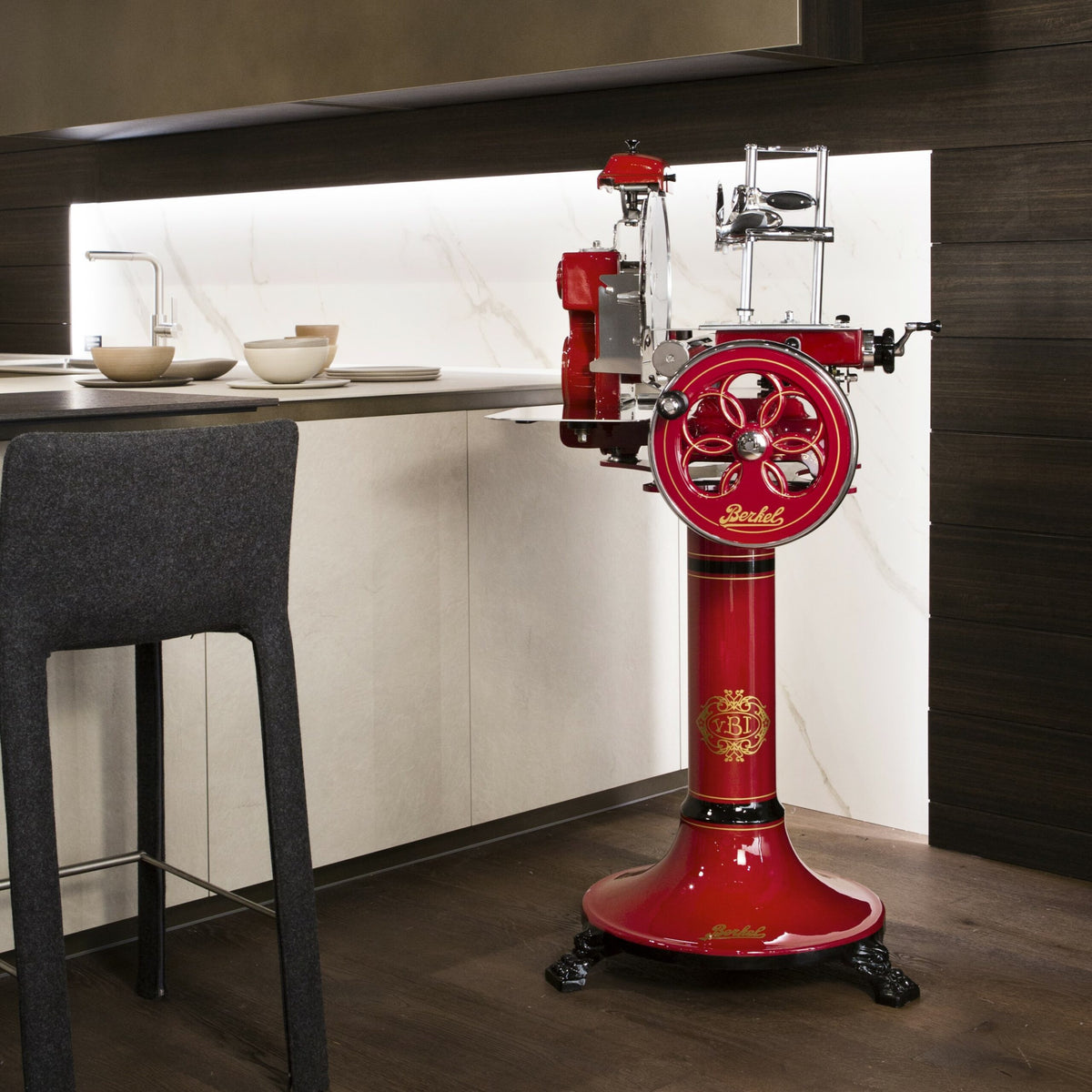 Eine elegante, moderne Küche mit weißen Marmorarbeitsplatten und dunklen Holzschränken, mit einer rot-chromfarbenen Berkel Volano Tribute Rot Aufschnittmaschine im Vintage-Stil mit floralem Schwungrad auf einem Sockel und einem Fassholz-Schneidebrett an der Theke. Die Küchentheke hat zwei Spülbecken und steht neben einem dunkelgrauen gepolsterten Stuhl.