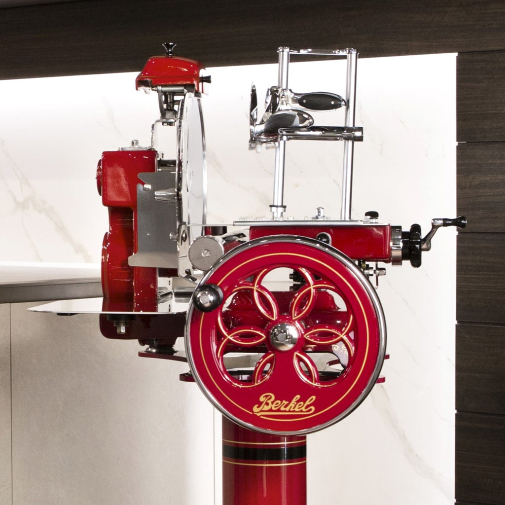 Eine Berkel Volano Tribute Rot Aufschnittmaschine mit Blumenschwungrad und einem Aufschnittbrett aus Weinfassholz vor einem modernen Küchenhintergrund mit dunklen und hellen Kontrasten. Der Allesschneider verfügt über ein großes, kunstvoll detailliertes Blumenschwungrad und polierte Metallkomponenten, die eine Mischung aus klassischem und modernem Design widerspiegeln.