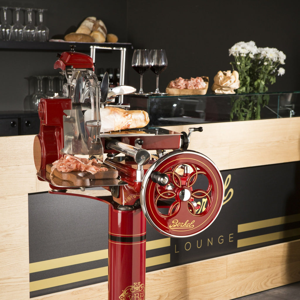 An einer modernen Feinkosttheke ist die Berkel Volano Tribute Rot Aufschnittmaschine mit Blumenschwungrad mit Standfuß + Aufschnittbrett aus Fassholz als eleganter Vintage-Aufschnittschneider für rotes Fleisch aufgestellt. Aufgeschnittener Schinken ruht elegant auf seinem Holzbrett, im Hintergrund sind Brot und Käse kunstvoll angeordnet. Zwei Gläser Rotwein und ein Strauß weißer Blumen ergänzen die einladende Präsentation auf der Theke.