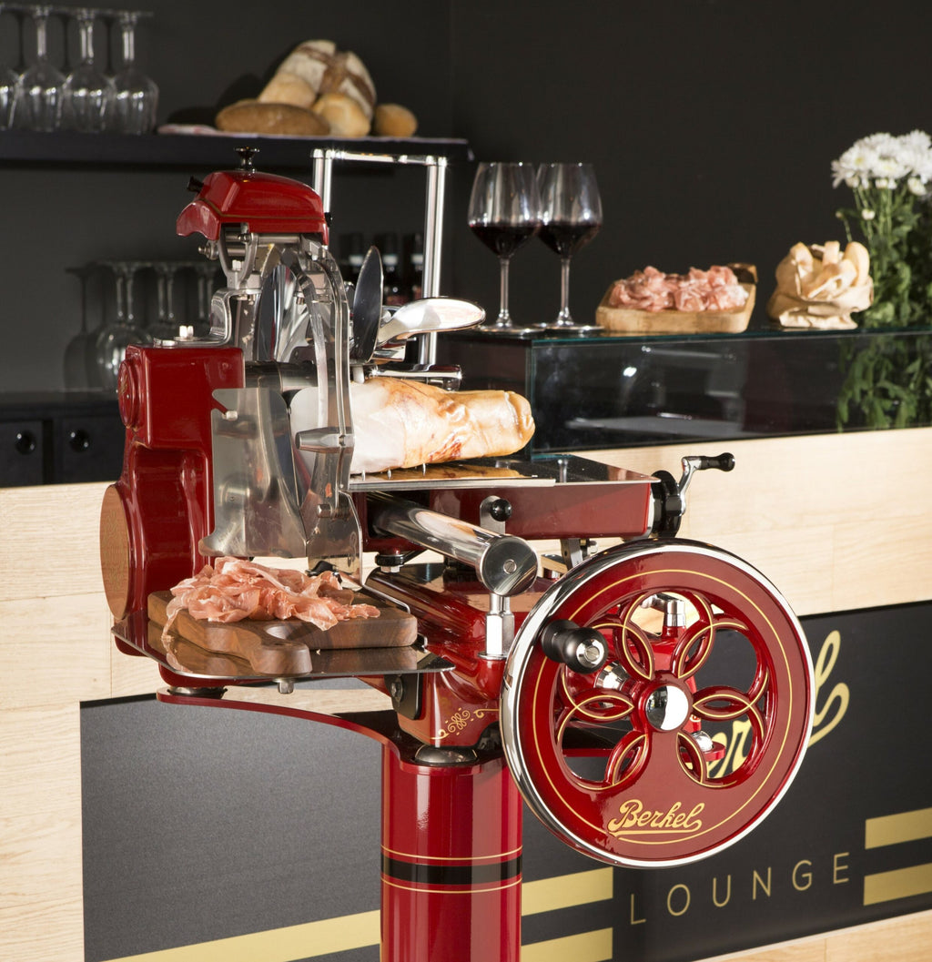 Eine alte Berkel Volano Tribute Rot Aufschnittmaschine mit Blumenschwungrad steht auf einer Holztheke aus Weinfassholz und schneidet sorgfältig Wurst. Dahinter befindet sich ein Barbereich mit Gläsern, Flaschen und zwei Gläsern Rotwein auf der Theke neben einem Laib Brot im Hintergrund.
