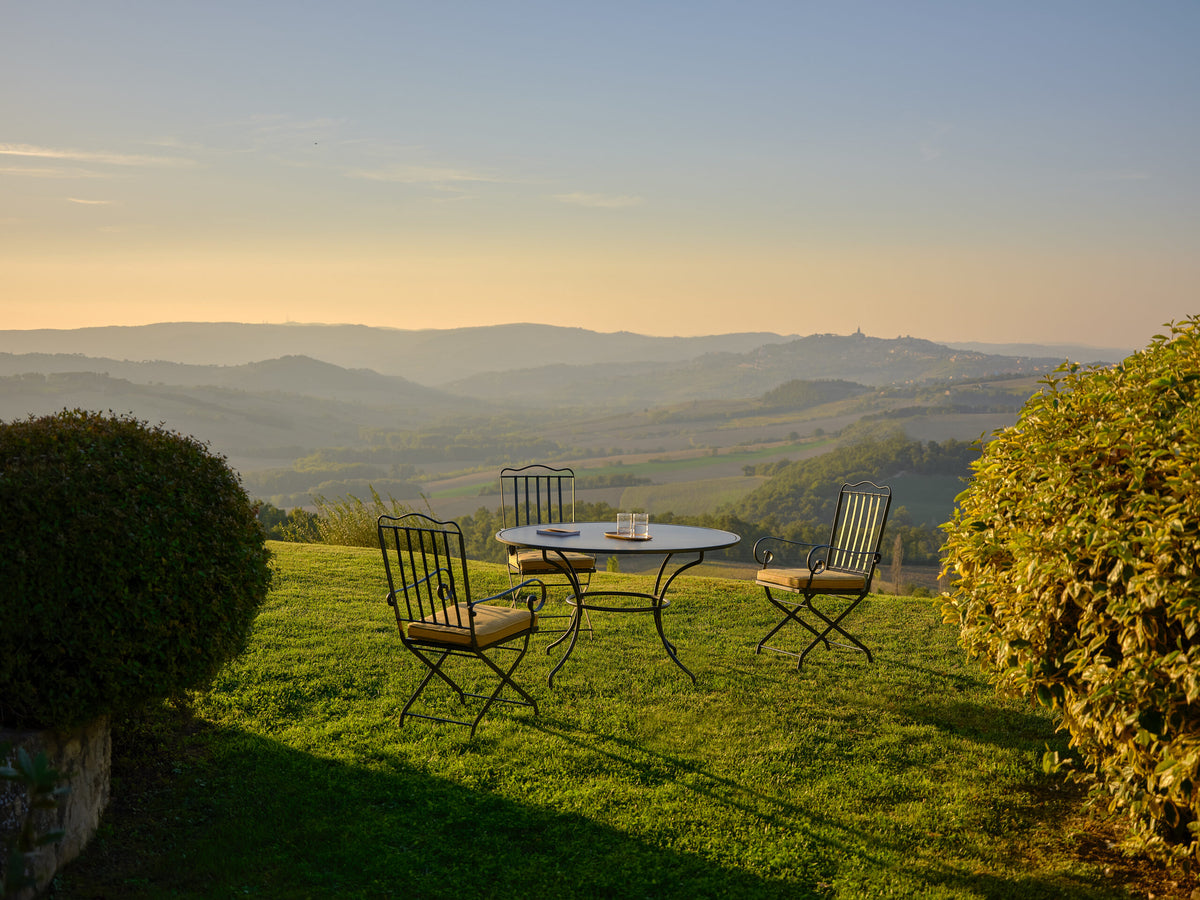 Eine Außeneinrichtung mit einem graphitgrauen Eisentisch und vier Sesseln Toscana aus Eisen in graphitgrau auf einer Rasenfläche mit Blick auf eine malerische Landschaft mit sanften Hügeln bei Sonnenuntergang. Auf dem Tisch stehen ein Buch und Kerzen, auf beiden Seiten von Büschen eingerahmt. Die Atmosphäre ist heiter und einladend.