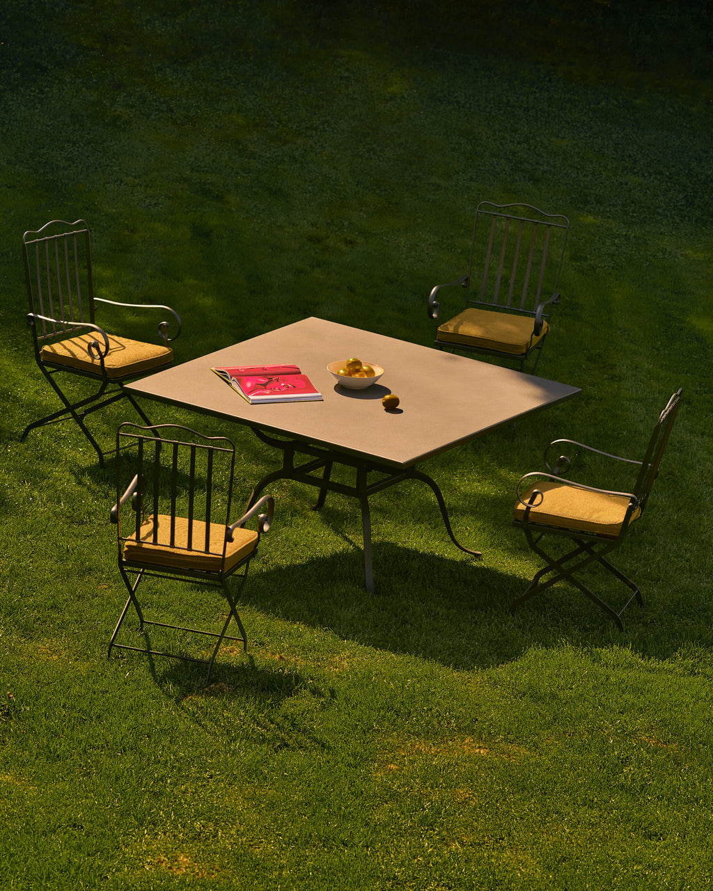 Ein quadratischer Gartentisch mit ein paar Gegenständen darauf steht auf einer Rasenfläche. Vier Sessel Toscana aus Eisen in graphitgrau, jeweils mit gepolsterten Sitzflächen, sind um den Tisch herum platziert und bilden einen gemütlichen Sitzbereich im Freien. Auf dem Tisch liegen ein rotes Buch und eine Schale mit gelbem Obst.