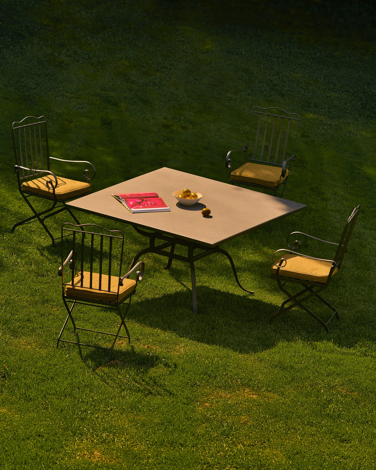 Ein quadratischer Gartentisch mit ein paar Gegenständen darauf steht auf einer Rasenfläche. Vier Sessel Toscana aus Eisen in graphitgrau, jeweils mit gepolsterten Sitzflächen, sind um den Tisch herum platziert und bilden einen gemütlichen Sitzbereich im Freien. Auf dem Tisch liegen ein rotes Buch und eine Schale mit gelbem Obst.