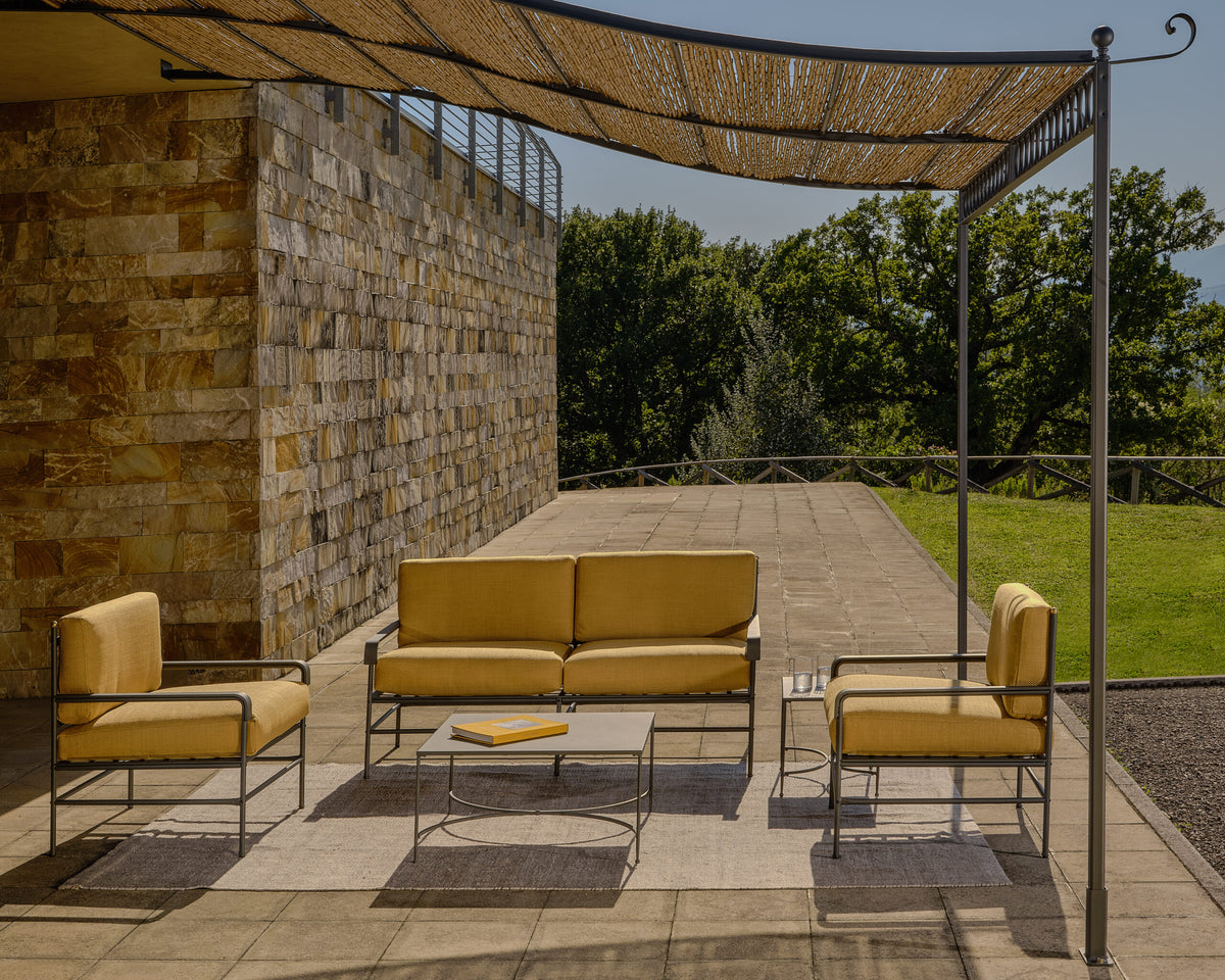 Eine moderne Terrasseneinrichtung mit Möbeln mit Metallrahmen und gelben Kissen unter einem geflochtenen Baldachin. Das Set umfasst ein Sofa, zwei Sessel und einen Couchtisch auf einer gefliesten Terrasse mit Teppichen. Eine Anbaupergola Tibisco mit Überdachung aus Polykarbonat überdeckt den Raum, während im Hintergrund ein Steingebäude und Grünflächen zu sehen sind.
