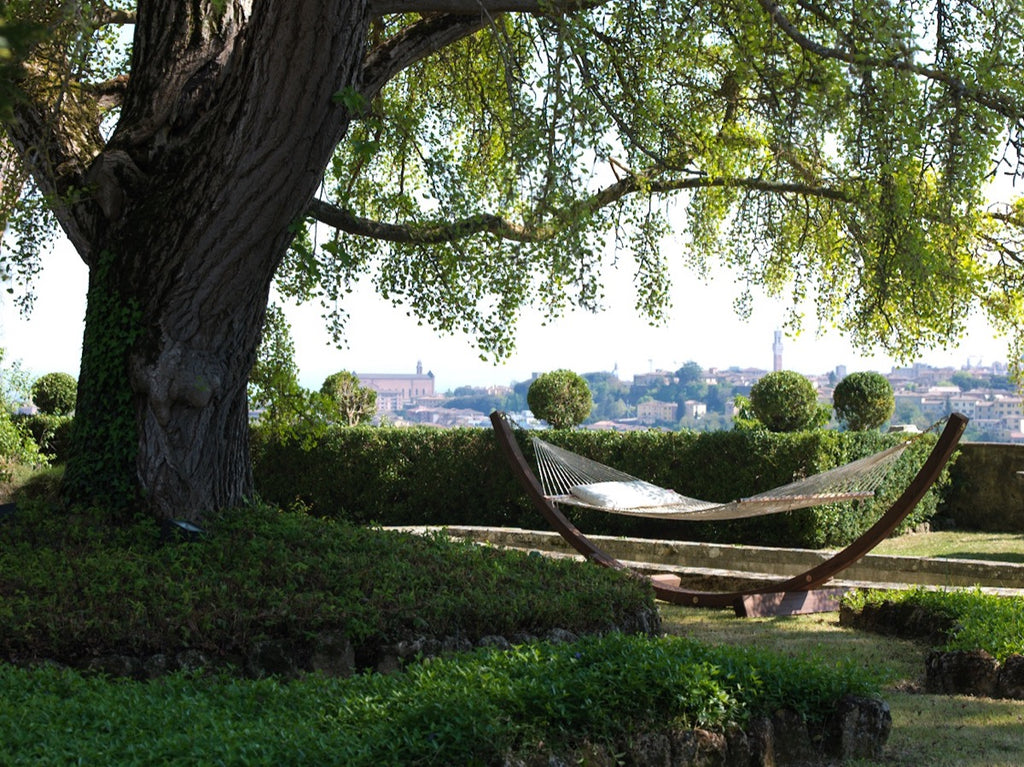 Eine Hängematte mit Unterteil aus verzinktem Stahl hängt zwischen einem großen Baum und einem Holzrahmen in einem üppigen Garten und bietet einen ruhigen Ruheplatz. Der Garten ist voller grüner Büsche und Gras, vor dem Hintergrund einer entfernten Stadt mit verschiedenen Gebäuden und einem blauen Himmel.
