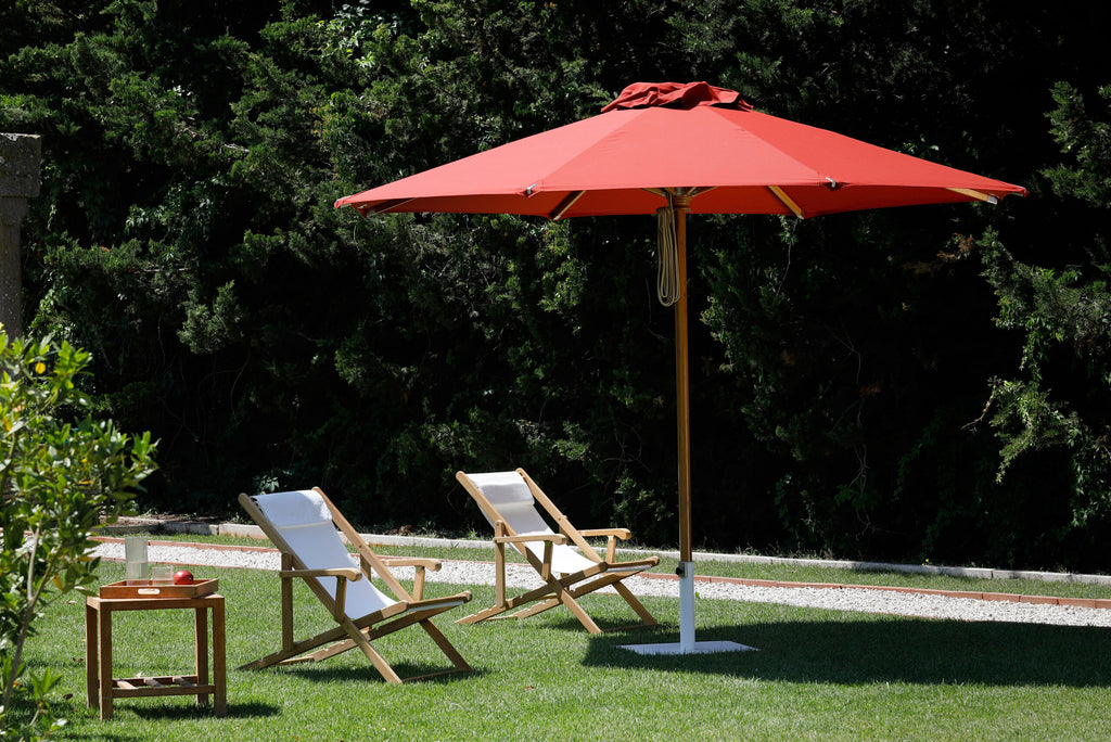 Zwei hölzerne Loungesessel mit weißen Kissen stehen unter einem großen roten Sonnenschirm Lipari (350 - 400 cm) in einem Garten. Auf einem kleinen Holztisch steht ein rotes Tablett mit einem Gegenstand darauf. Im Hintergrund sind üppige grüne Bäume und Büsche zu sehen, die eine perfekte ruhige Atmosphäre schaffen.