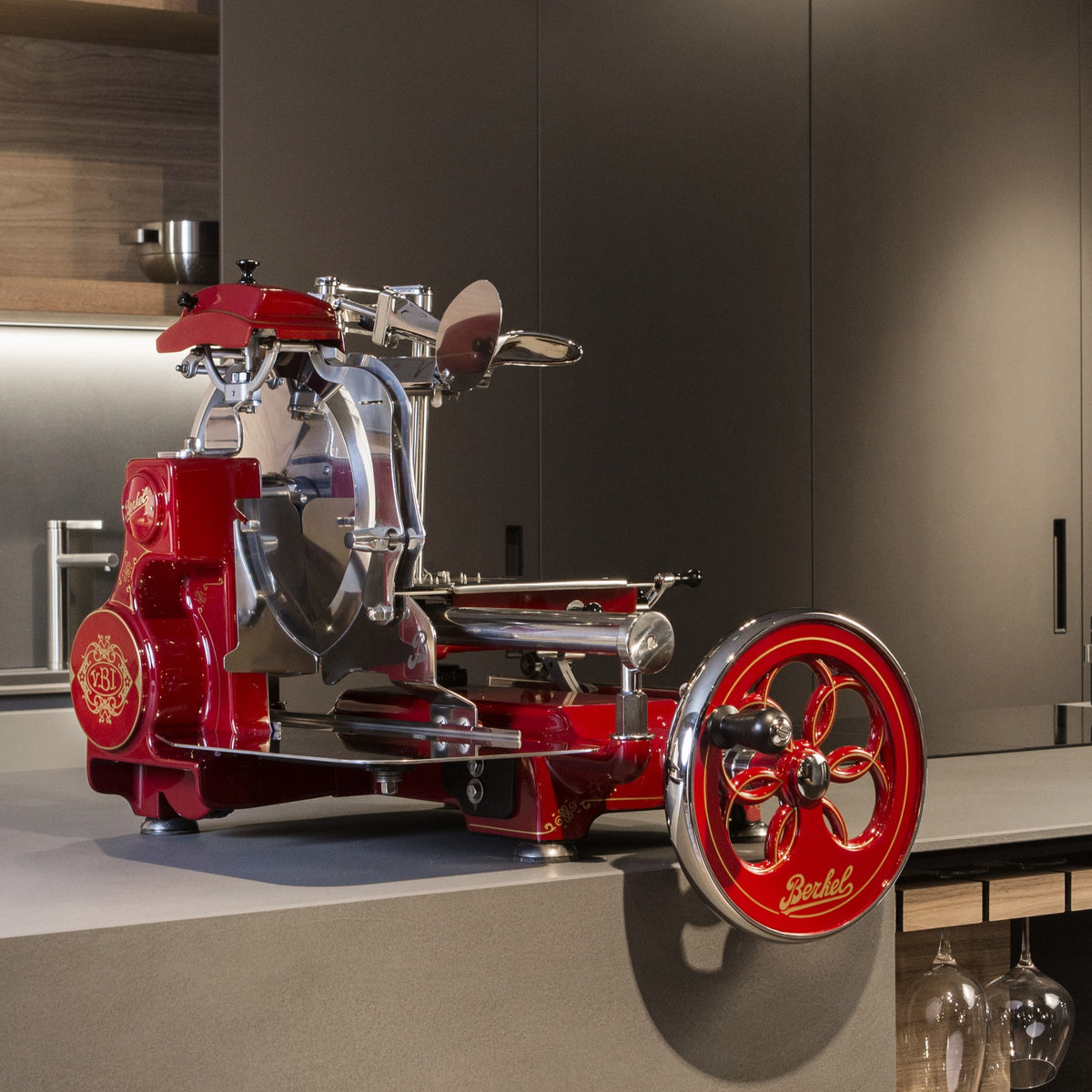 Eine rot-chromfarbene Berkel Volano Tribute Rot Aufschnittmaschine im Vintage-Stil mit floralem Handkurbelrad und Fassholz-Schneidebrettständer steht auf einer Küchentheke. Die Aufschnittmaschine ist mit aufwendigen Details versehen und der Markenname „Berkel“ ist deutlich zu sehen. Die moderne Küchenkulisse besteht aus Holzschränken und minimalistischem Dekor.