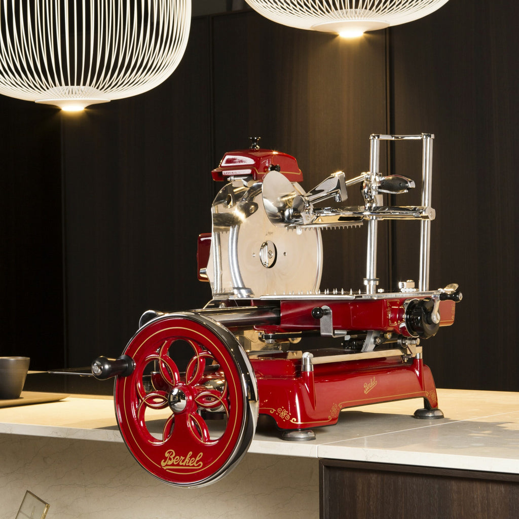 Eine Berkel Volano Tribute Rot Aufschnittmaschine mit Blumenschwungrad, komplett mit Ständer und Schneidebrett aus Fassholz, steht auf einer Marmorarbeitsplatte. Die Aufschnittmaschine im Vintage-Stil in Rot und Chrom verfügt über ein großes dekoratives Handkurbelrad und komplizierte Metallkomponenten, vor einem Hintergrund aus dunklen Schränken und beleuchtet von zwei modernen Hängelampen.