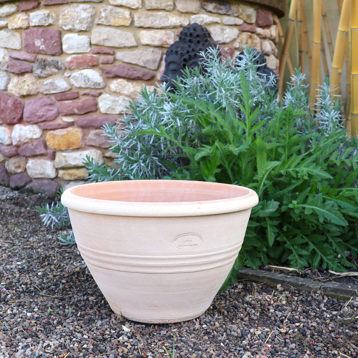 Ein großer, leerer frostfester Blumentopf, Blumenkübel „Veronica“ D=35 cm steht auf einer Kiesfläche in einem Garten. Dahinter befinden sich grüne, blattreiche Pflanzen und ein Abschnitt einer Steinmauer mit verschiedenen Farben. Auf der rechten Seite sind einige Bambusstäbe und wunderschöne Veronica-Blüten zu sehen. Die Szene ist im Freien und natürlich.