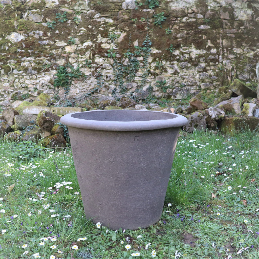 Das große, leere Pflanzgefäß „Atelier Vierkant“, H=65 cm, steht auf Gras mit kleinen weißen Blüten. Im Hintergrund ist eine Steinmauer zu sehen, die teilweise mit grünem Efeu bewachsen ist.