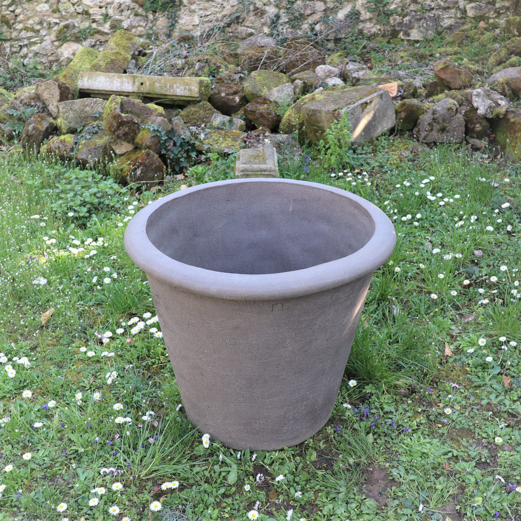 Ein großer, grauer Blumentopf „Atelier Vierkant“ mit einer Höhe von 65 cm ist leer und steht mit kleinen weißen Blumen auf Gras, vor der Kulisse einer Steinmauer und eines Steinhaufens.