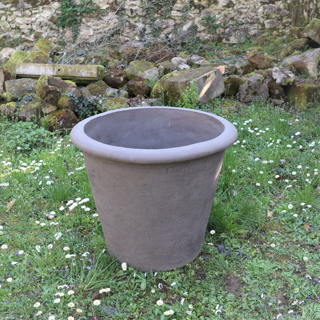 Ein großer, leerer grauer Pflanztopf namens Pflanzgefäß „Atelier Vierkant“, H=65 cm, steht auf mit kleinen weißen Blumen geschmücktem Gras, vor dem Hintergrund einer Steinmauer und moosbedeckter Felsen, und schafft so eine heitere Atmosphäre.