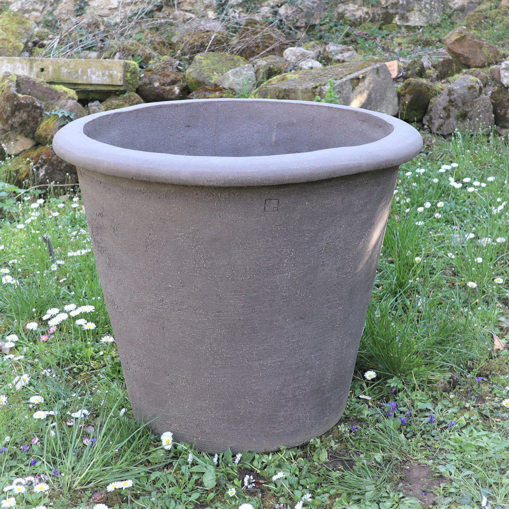 Ein großes, graues Pflanzgefäß „Atelier Vierkant“, H=65 cm, mit breitem Rand, steht im Gras und ist von kleinen weißen und violetten Blüten umgeben. Felsen und eine Steinmauer im Hintergrund unterstreichen seine Statur.