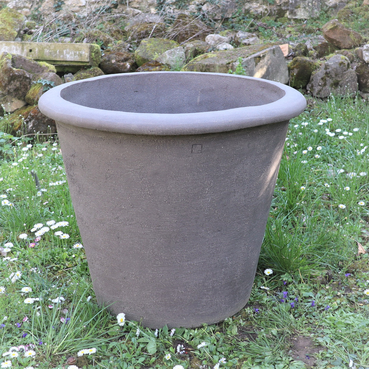Ein großes, graues Pflanzgefäß „Atelier Vierkant“, H=65 cm, mit breitem Rand, steht im Gras und ist von kleinen weißen und violetten Blüten umgeben. Felsen und eine Steinmauer im Hintergrund unterstreichen seine Statur.