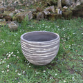 Ein großes, rundes Pflanzgefäß „Atelier Vierkant“, H=50, mit Streifen steht auf der Wiese inmitten kleiner weißer Blumen, während moosbewachsene Felsen und eine Steinmauer eine malerische Kulisse bilden.