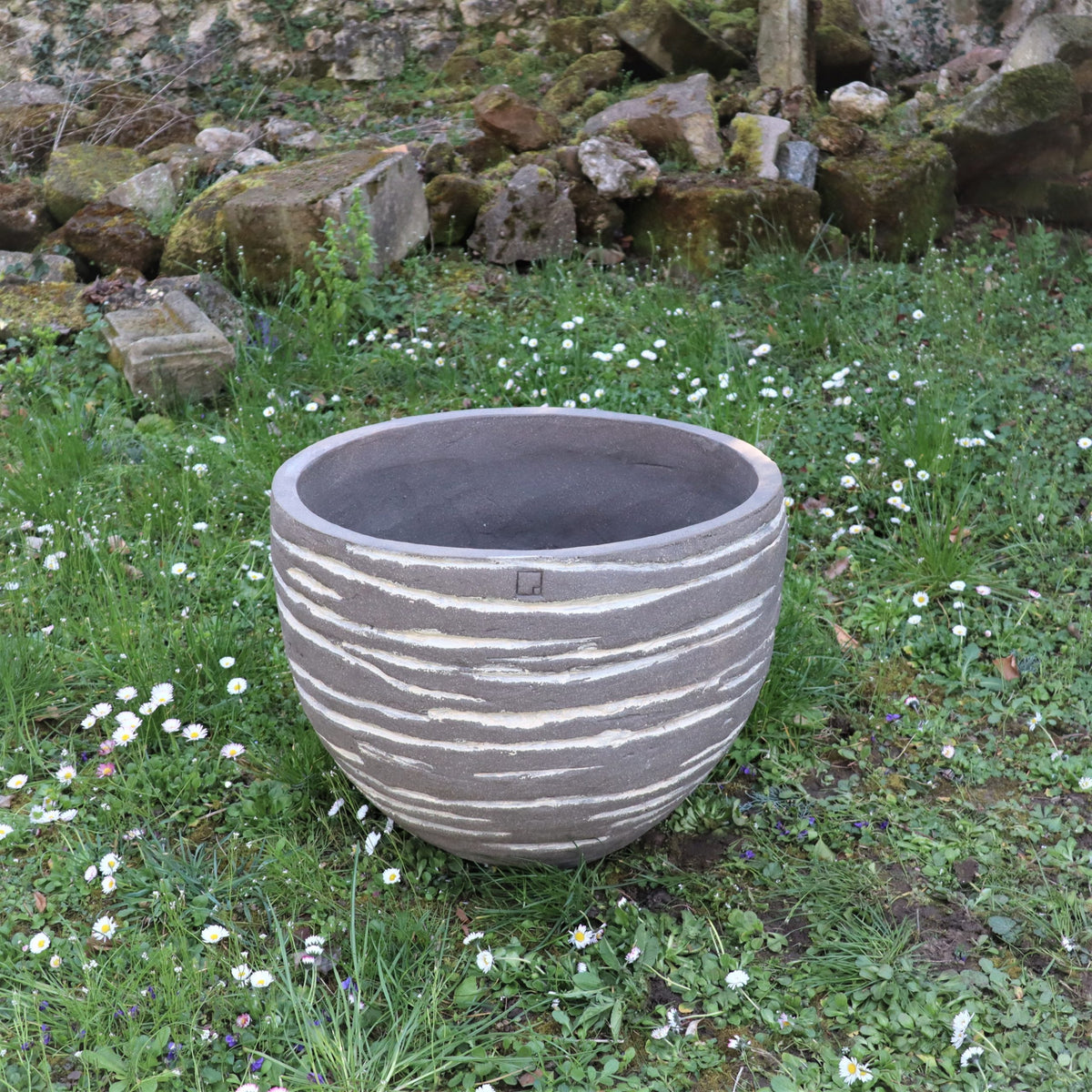 Ein großes, rundes Pflanzgefäß „Atelier Vierkant“, H=50, mit Streifen steht auf der Wiese inmitten kleiner weißer Blumen, während moosbewachsene Felsen und eine Steinmauer eine malerische Kulisse bilden.