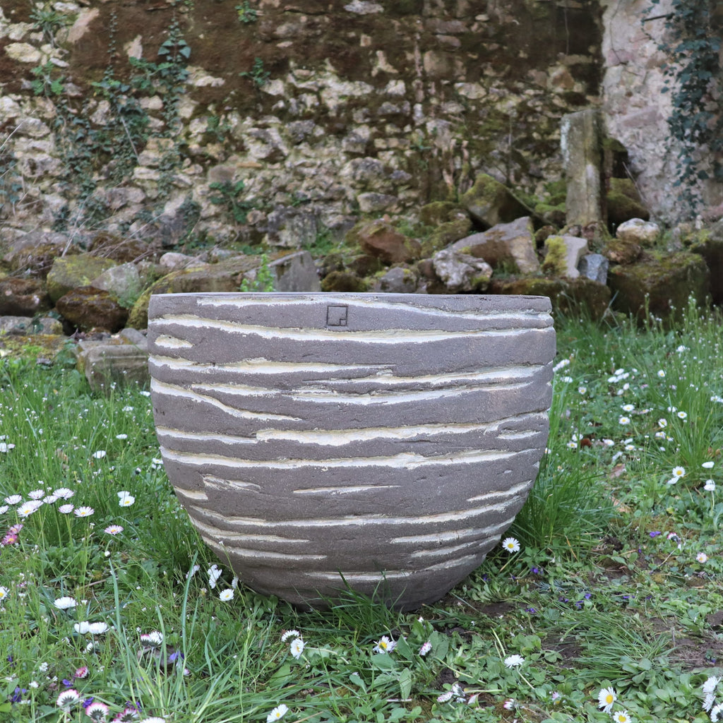 Ein großes strukturiertes Pflanzgefäß „Atelier Vierkant“, H=50, mit Streifen, steht auf Gras mit kleinen weißen Blüten, vor einer verwitterten, teilweise mit Efeu bewachsenen Steinmauer.