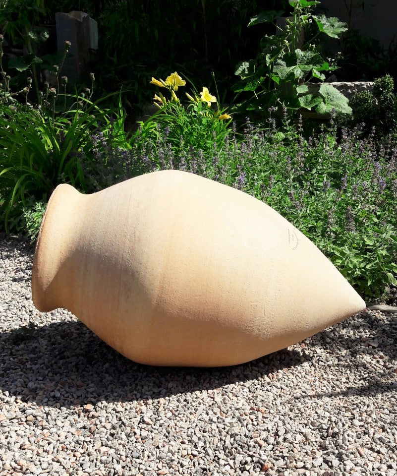 Die liegende Spitzamphore "Vitex 2", H=60cm, ruht auf Kies inmitten von Grünpflanzen und gelben Blüten in einem sonnenbeschienenen Garten.
