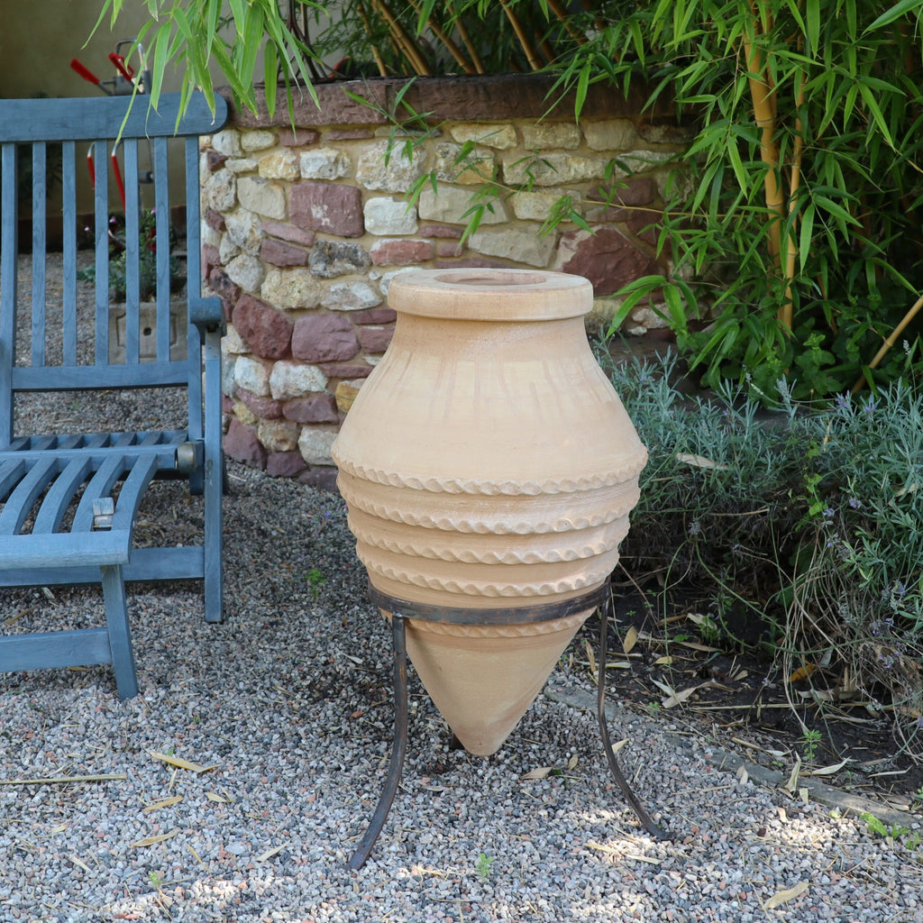 Eine große, dekorative, liegende Amphore „Vitex“ mit spitzem Fuß, 60 cm hoch und auf dem dazugehörigen Metallständer ruhend, schmückt eine Gartenumgebung im Freien. Sie steht neben einem blauen Holzstuhl inmitten üppigen Grüns, der Boden ist mit Kies bedeckt.