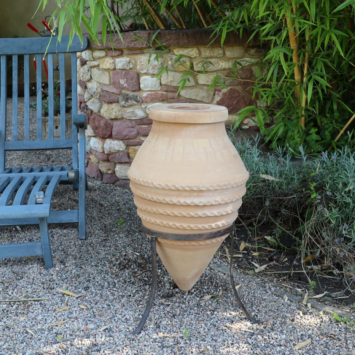 Eine große, dekorative, liegende Amphore „Vitex“ mit spitzem Fuß, 60 cm hoch und auf dem dazugehörigen Metallständer ruhend, schmückt eine Gartenumgebung im Freien. Sie steht neben einem blauen Holzstuhl inmitten üppigen Grüns, der Boden ist mit Kies bedeckt.