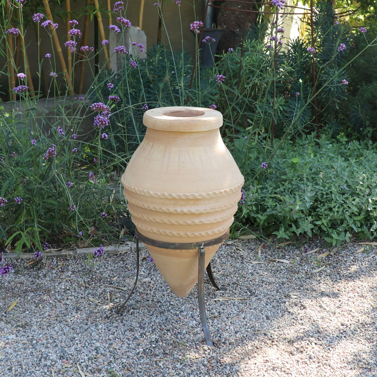 Die liegende Amphore „Vitex“, eine große beige Keramikurne mit dekorativen Rillen, steht auf einem Metallständer und ist im Freien in einer Gartenumgebung platziert. Um die Urne herum stehen saftige grüne Pflanzen und violette Vitex-Blüten, während kleine Kieselsteine den Boden bedecken.