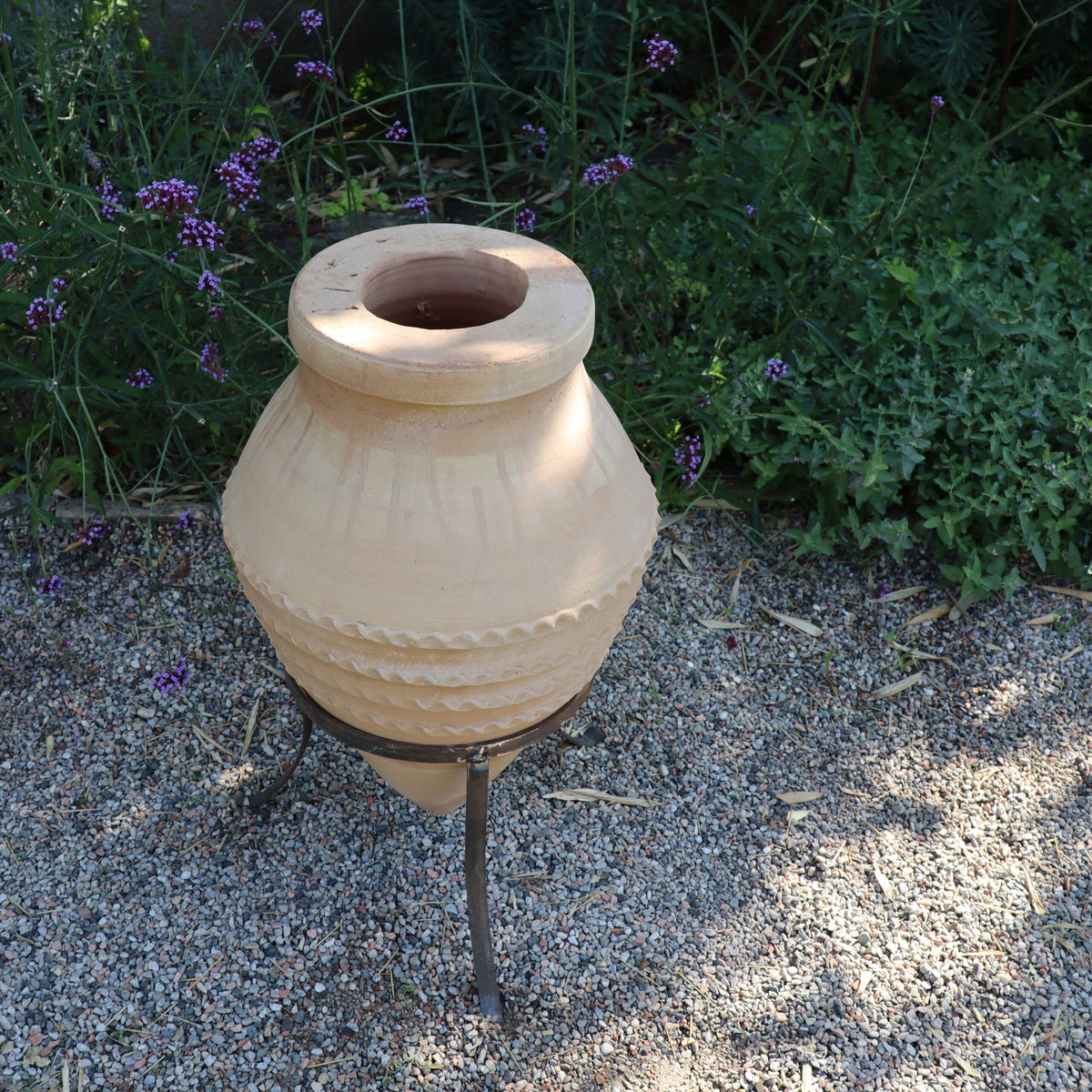 Ein Bild der liegenden Amphore „Vitex“, einer großen, beigefarbenen Keramikamphore mit komplizierten Mustern, die auf einem Metallständer steht. Die Amphore steht auf einer Kiesfläche, umgeben von üppigen grünen Pflanzen und violetten Vitex-Blüten. Sonnenlicht wirft Schatten und hebt die Textur der Vase hervor.