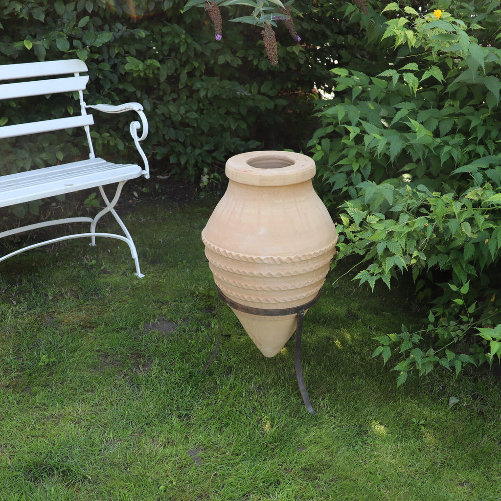 Die liegende Amphore „Vitex“, H=60 cm mit Ständer, schmückt den Garten. Ihre große beige Form weist dekorative horizontale Rillen auf und thront elegant auf einem Metallständer. Daneben steht eine weiße Metallbank mit aufwendig gestalteten Armlehnen, die vor üppigem grünem Laub steht, auf dem die Vitex-Blüten wunderschön blühen.