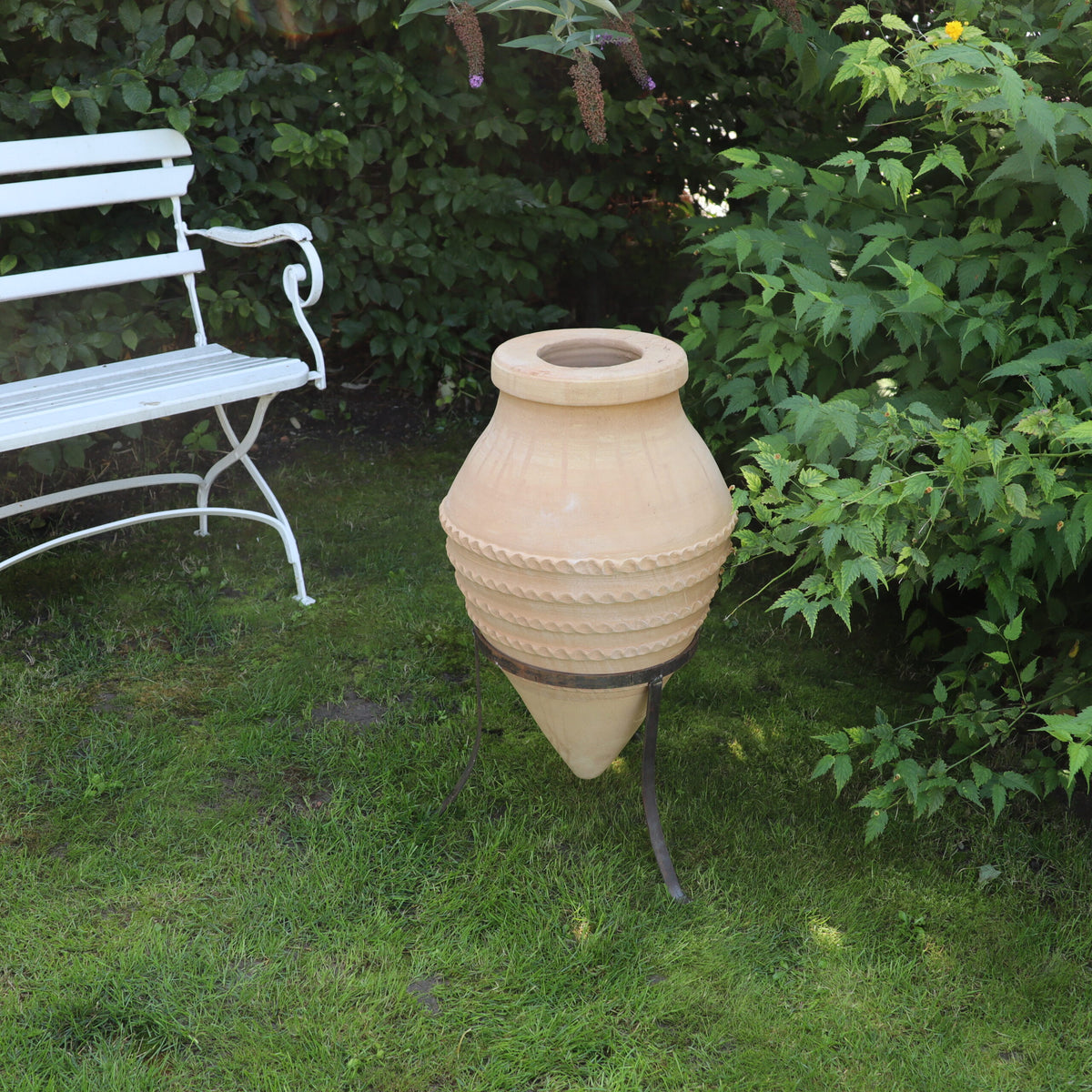 Die liegende Amphore „Vitex“, H=60 cm mit Ständer, schmückt den Garten. Ihre große beige Form weist dekorative horizontale Rillen auf und thront elegant auf einem Metallständer. Daneben steht eine weiße Metallbank mit aufwendig gestalteten Armlehnen, die vor üppigem grünem Laub steht, auf dem die Vitex-Blüten wunderschön blühen.