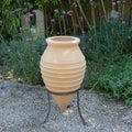 Die ruhige Gartenszene zeigt die liegende Amphore „Vitex“, die 60 cm hoch ist und einen Metallständer hat. Dieser Terrakottatopf, der mit horizontalen Rillen verziert ist und sich nach unten verjüngt, ist von grünen Bambuspflanzen und violetten Vitex-Büschen umgeben. Kleine Kieselsteine bedecken den Boden und verstärken den natürlichen Charme.