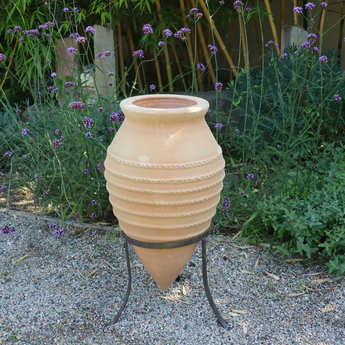 Die ruhige Gartenszene zeigt die liegende Amphore „Vitex“, die 60 cm hoch ist und einen Metallständer hat. Dieser Terrakottatopf, der mit horizontalen Rillen verziert ist und sich nach unten verjüngt, ist von grünen Bambuspflanzen und violetten Vitex-Büschen umgeben. Kleine Kieselsteine bedecken den Boden und verstärken den natürlichen Charme.