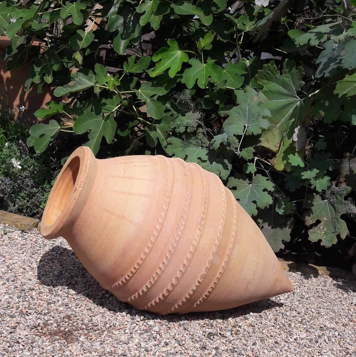Eine große, beigefarbene Amphore aus Keramik oder Ton aus der handgefertigten liegenden Amphoren-Kollektion „Vitex“ mit einer Höhe von 70 cm liegt seitlich auf einer Kiesfläche. Sie ist mit erhabenen kreisförmigen Mustern verziert und steht vor einem Hintergrund aus üppigem grünem Laub.