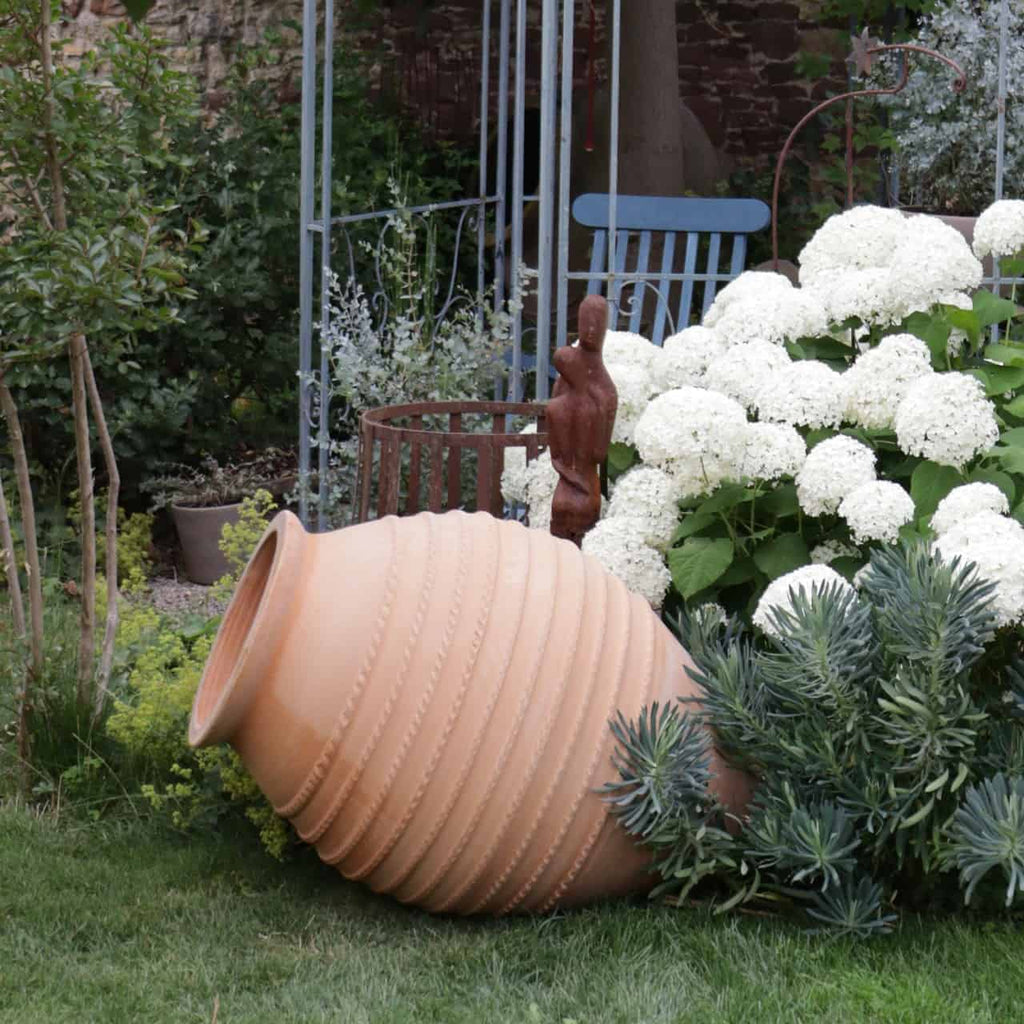 Eine große, gerippte Terrakotta-Urne, die Große liegende Amphore „Vitex“ (H=1,1 m), liegt auf der Seite in einem Garten, umgeben von üppigem Grün und weißen Hortensienblüten. Hinter der Urne steht eine bronzene Statue einer stehenden menschlichen Figur (H=1 m). Im Hintergrund sind ein blauer Gartenstuhl und ein Metallgitter zu sehen.