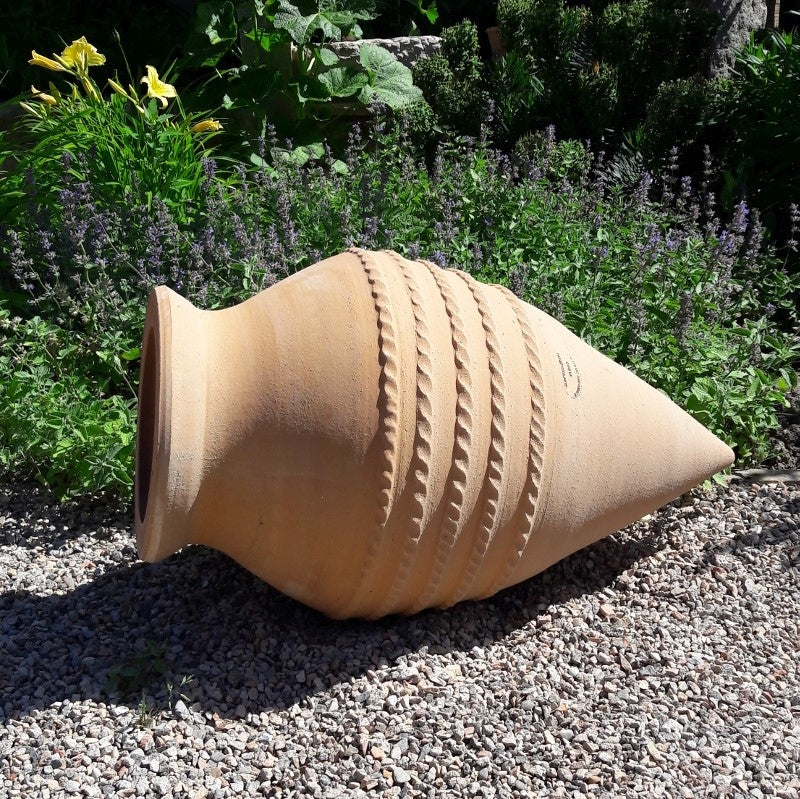Die Liegende Amphore "Vitex", H=60cm, ist eine dekorative Terrakotta-Urne mit strukturierten Bändern, die auf der Seite liegend an einem Kiesweg und umgeben von üppigem Grün und blühenden Blumen in einem Garten steht.