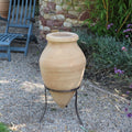 Die handgefertigte Terrakotta Spitzamphore „Vitex 2“ mit Metallständer 60 cm steht auf einem schwarzen Metallständer in einer Gartenkulisse. Um sie herum befinden sich Kiesboden, Grünpflanzen und im Hintergrund eine Steinmauer. Links steht ein blauer Holzstuhl. Im Hintergrund rechts sind violette Blumen zu sehen.
