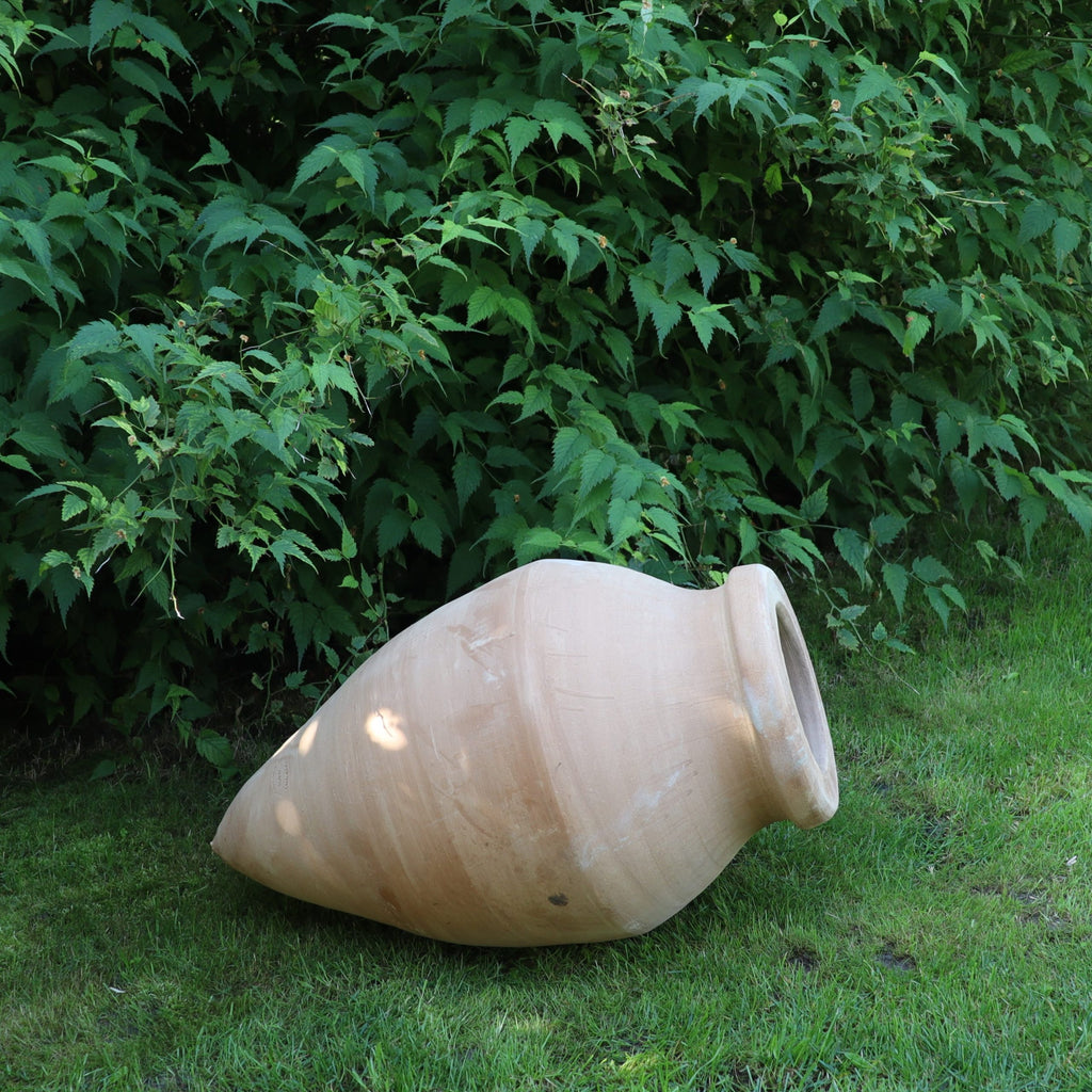 Die handgefertigte Terracotta Spitzamphore "Vitex 2" mit Metallständer (60 cm) liegt seitlich auf grünem Gras vor dichten Büschen und eignet sich perfekt als stilvolles Garten-Highlight.