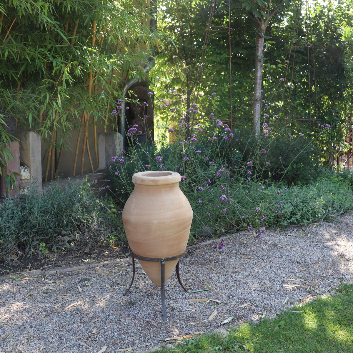 Eine handgefertigte Terrakotta-Spitzamphore „Vitex 2“ mit Metallständer 60 cm steht stolz im Garten. Der Garten ist mit grünem Blattwerk, darunter breitblättrige Pflanzen und Büsche, ausgestattet. Kieswege schlängeln sich um die Vegetation und schaffen eine ruhige, geordnete natürliche Umgebung.