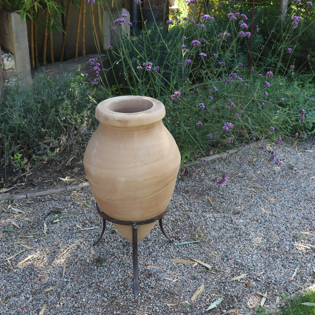 Die handgefertigte Terrakotta-Spitzamphore „Vitex 2“ mit Metallständer 60 cm steht auf einem Kiesweg in einem Garten. Der Topf ist von üppigen grünen Pflanzen und violetten Vitex 2-Blüten umgeben, die eine ruhige und natürliche Atmosphäre schaffen.