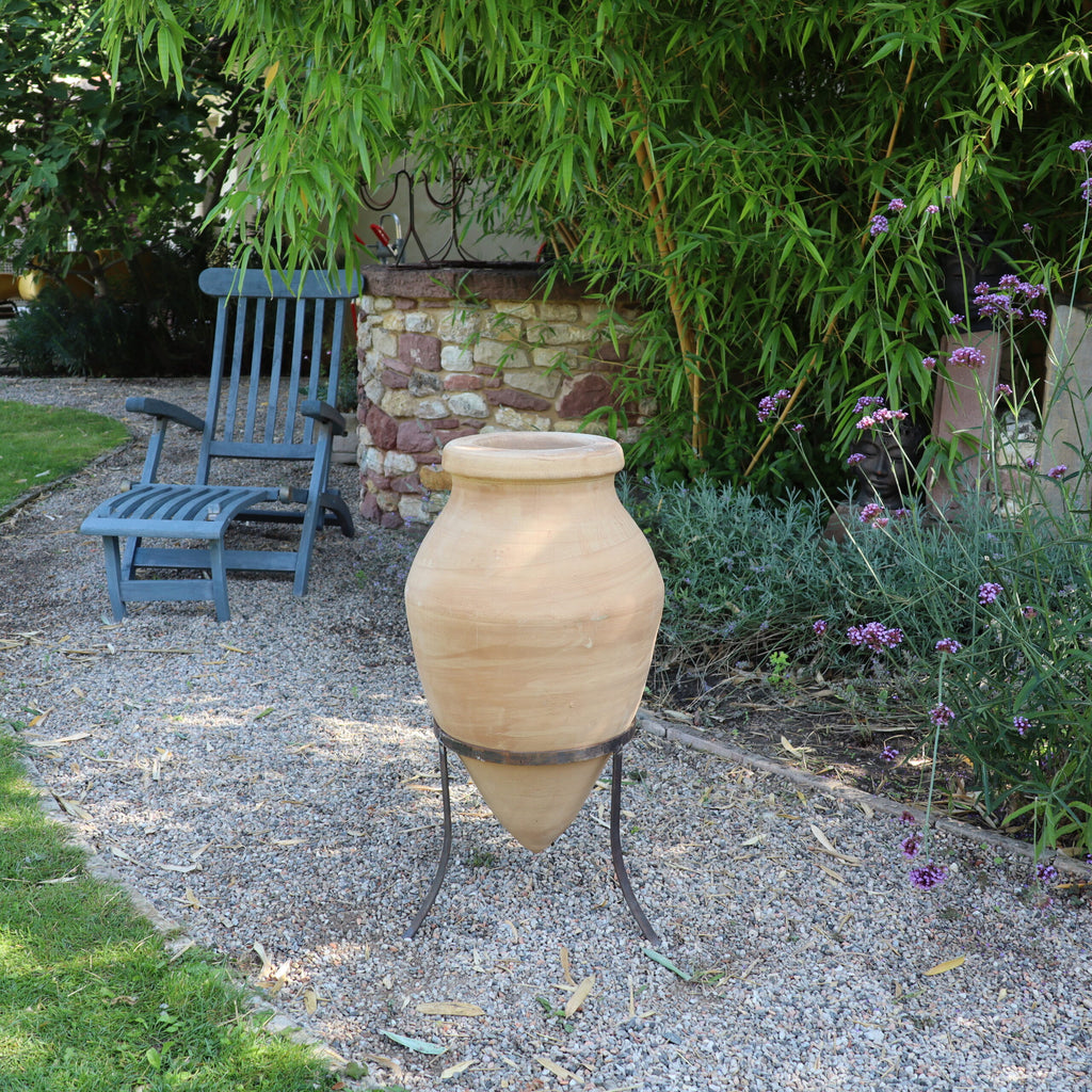 Eine große, handgefertigte Terrakotta-Spitzamphore „Vitex 2“ mit Metallständer (60 cm) steht auf einem Metallständer entlang eines Kieswegs im Garten. Links davon steht ein blauer Holzstuhl neben einer Steinmauer. Der Garten ist üppig grün, darunter hoher Bambus und violette Blumen.