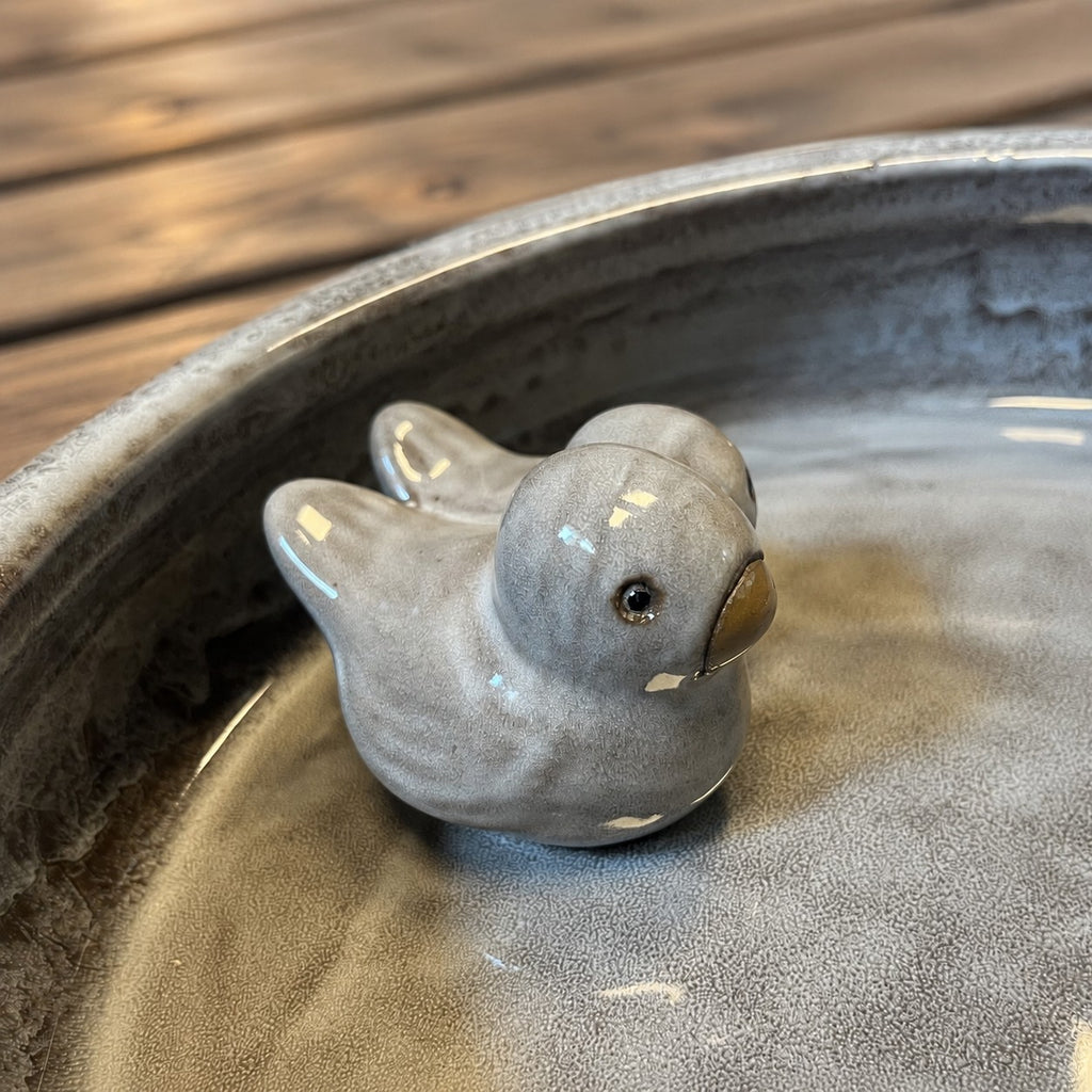 Ein kleiner, glänzender, grauer Keramikvogel sitzt in einer runden, flachen, grauen Schale, im Hintergrund ist eine Holzoberfläche zu sehen.