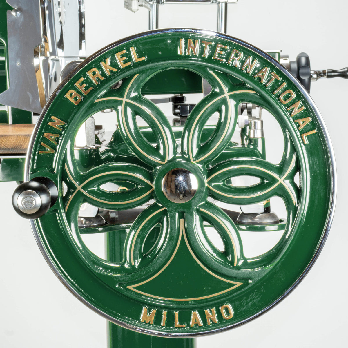Nahaufnahme des Vorderrads einer Berkel Volano B114-Fleischschneidemaschine in Sonderlackierung British Racing Green. Die verzierten Ausschnitte auf dem Rad zeigen den goldenen Text „VAN BERKEL INTERNATIONAL MILANO“. Im Hintergrund sind der Griff und andere Teile der grünen Vintage-Schneidemaschine zu sehen.