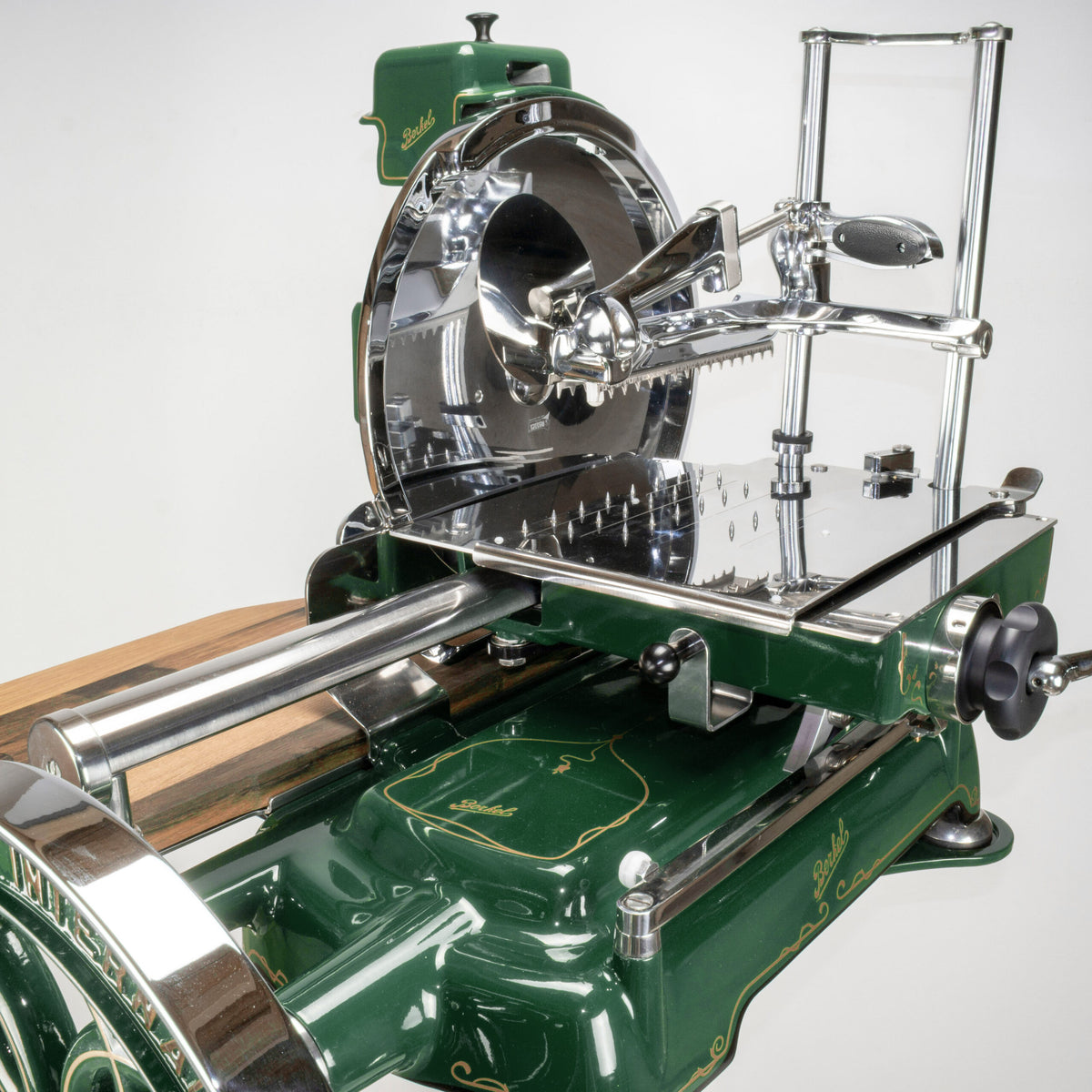 Eine Detailabbildung des Berkel Volano B114 in Sonderlackierung „British Racing Green“, einer Fleischschneidemaschine im Vintage-Stil mit Handkurbel, runder Klinge, Klemmmechanismus zum Halten des Fleisches und reich gestaltetem Sockel mit Silber- und Holzakzenten.
