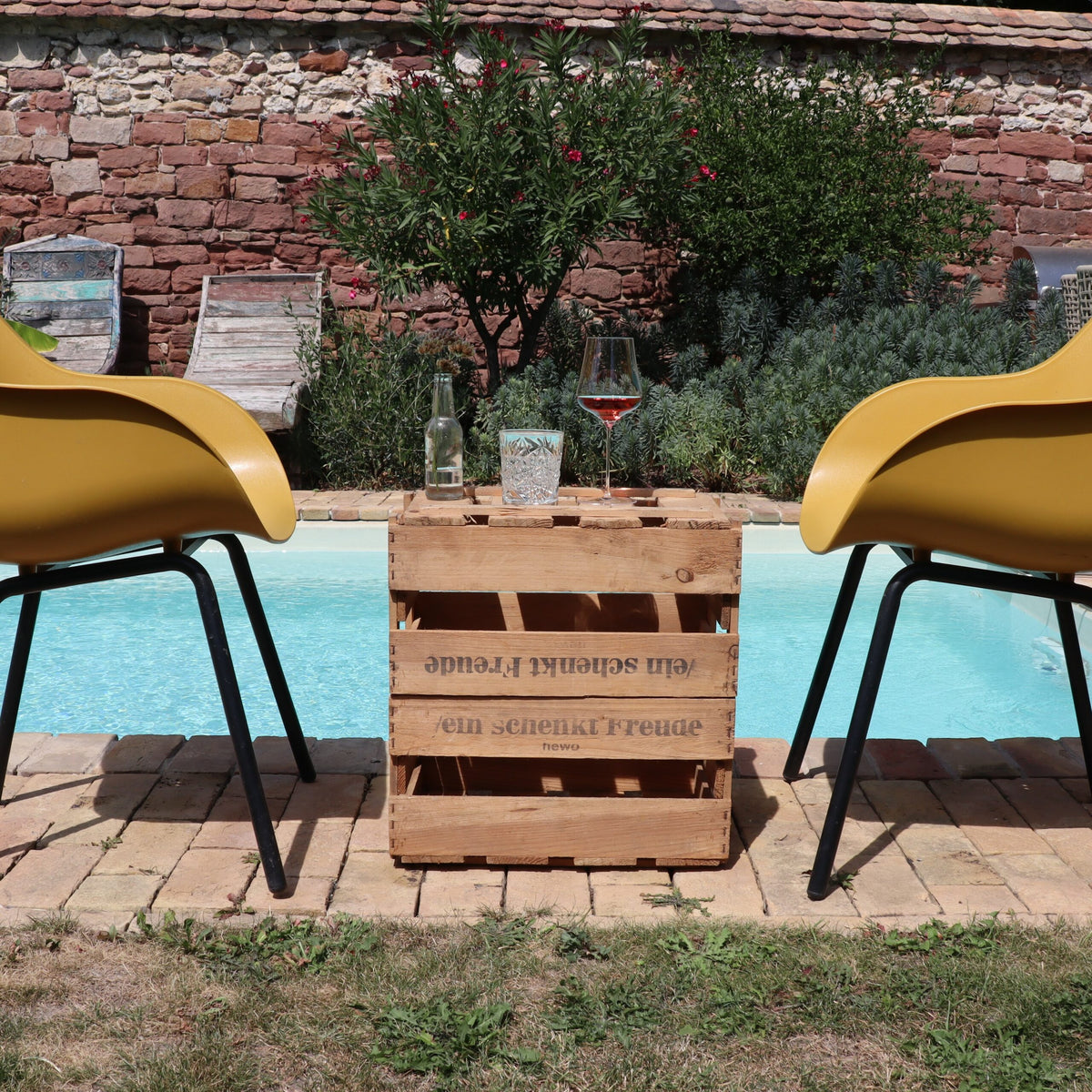 Zwei gelbe Stühle stehen vor einem Pool, dazwischen steht ein Tisch „Alte Weinkiste Holz Vintage aus der Pfalz mit Aufschrift“. Auf dem Tisch stehen eine Flasche Weißwein, ein mit Wasser gefülltes Glas und ein Glas Roséwein. Hinter dem Pool befindet sich ein üppiger Garten mit verschiedenen Pflanzen und einer roten Backsteinmauer.